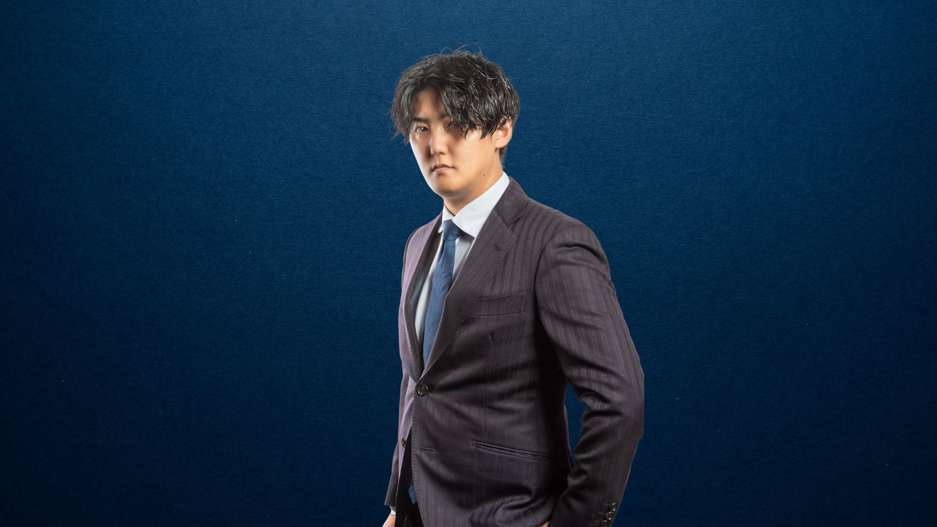 スーツを着た男性が正面を見つめて立っている写真（濃紺背景）